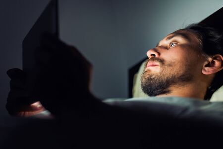 Young handsome and tired man with a beard cannot sleep and is watching something on his tablet at night.の写真素材