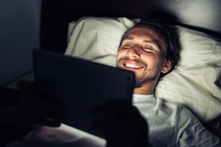 Handsome bearded smiling caucasian male lying in bed at night while watching something on his tablet.の写真素材