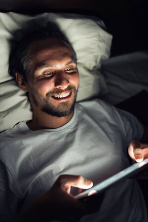 Handsome bearded smiling caucasian male lying in bed at night while watching something on his tablet.の写真素材