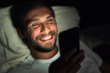 Bearded happy young man is lying in his bed and smiling while watching something on his phone. It is clearly very late at night.の写真素材