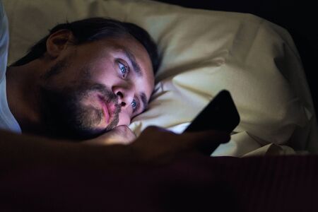 Young handsome and tired man with a beard cannot sleep and is watching something on his phone at night.の写真素材