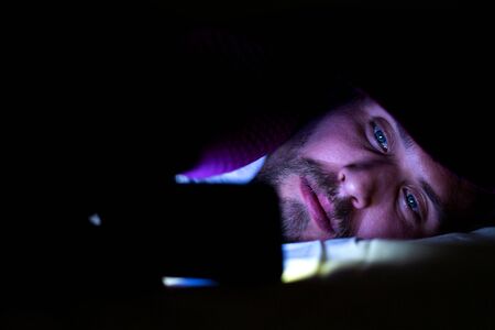 Bearded young man is lying in bed under his blanket. He cannot sleep and is watching something on his mobile phone.の写真素材