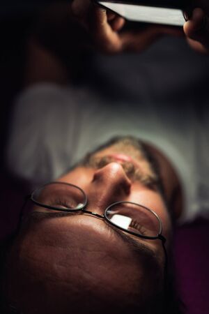 Bearded young man is lying in his bed at night while watching something on his mobile phone. Phone's reflection is visible in man's glasses.の写真素材