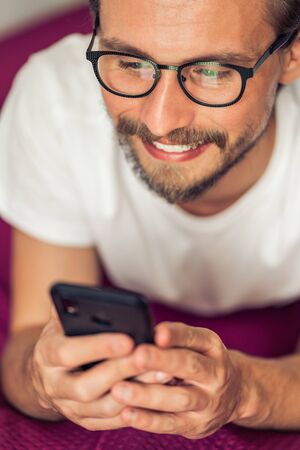 Handsome smiling young man with glasses lying on bed while holding his mobile phone.