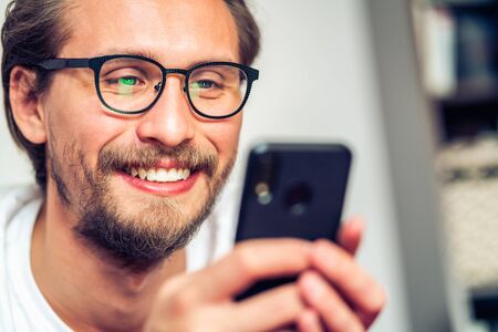 Handsome smiling young man with glasses lying on bed while holding his mobile phone.の写真素材