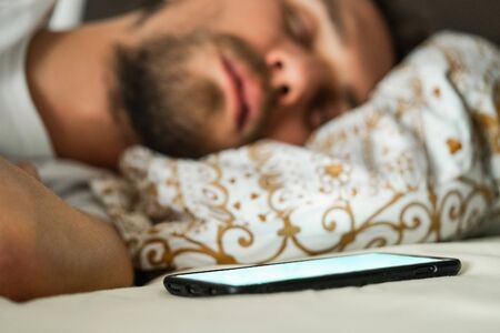 Close-up photo of a young man sleeping in bed. Mobile phone with a white blinking screen is visible in the foreground.の写真素材