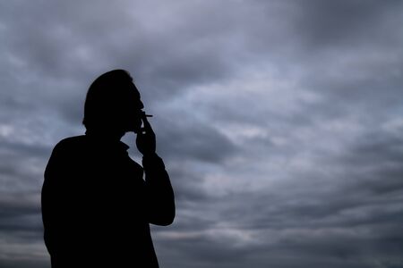 Dark silhouette of a handsome young man smoking a cigarette. Dark clouds visible in the background.