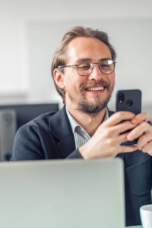 Young handsome man in glasses is interacting with his mobile phone while being at work. Happiness...