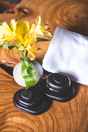 A view from above of yellow flowers in a vase and stones for stone therapy. Yellow daffodils and stones for massage lie on a wooden table. White towels lie in the background.の写真素材