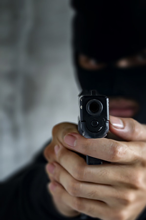 Masked robber with gun aiming into the camera against a black background. Selective focus.の写真素材