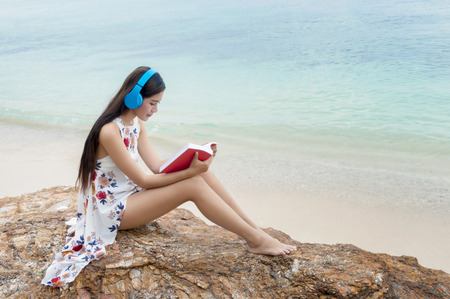 Beautiful smiling girl listening to music and reading a book on the beach of blue ocean and sky on the background.Relaxing concept.の写真素材