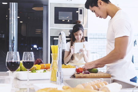 Beautiful asian young couple loving smiling is looking to cooking in kitchen at home,Happy handsome man cooking vegetables and Happy woman play tablet.の写真素材