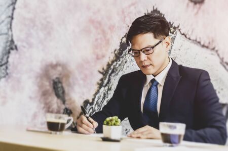 Confident businessman in eyeglasses looking at the paper and working at table with computer monitor in modern office.の写真素材