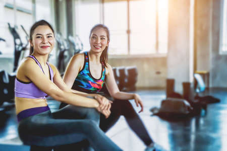 Two young asian fit is talking with her female friend something at gym. Both girls are resting after training together in sport center. They are sitting on floor and leaning on gym during.の写真素材