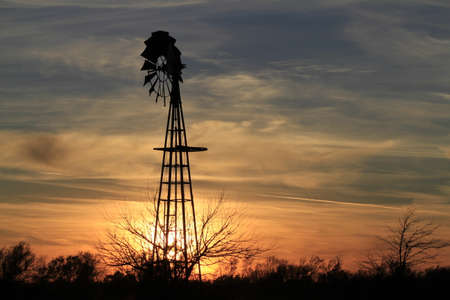 A shot of a Kansas Sunset with clouds, Tree's, and a Windmill silhouette north of Hutchinson Kansas USA out in the country.の写真素材