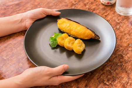 Grilled banana - Delicious golden brown grilled banana on black plate with mint leaves in woman's hand. Thai dessert. Traditional, Thai and Asian recipe menu.の写真素材