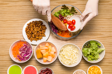 Fresh vegetable salad with insects - Human female hands cooking vegetable salad with worm insects in the kitchen on a wooden table. Healthy meal high protein diet concept. Top view. Selective focus.の写真素材