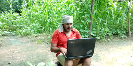DISTRICT KATNI, INDIA - AUGUST 14, 2019: An indian village farmer operating laptop around agriculture green field wearing common cloth on head.のeditorial素材