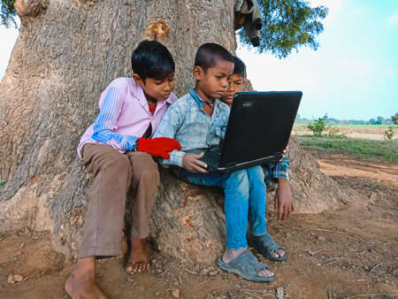 DISTRICT KATNI, INDIA - JANUARY 01, 2020: An indian boy teaching about laptop technology to the friends at open area green field near the tree.のeditorial素材