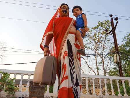 DISTRICT KATNI, INDIA - MAY 21, 2020: An indian village woman wearing face mask for corona virus protection with holded kid on lap at sky background.のeditorial素材