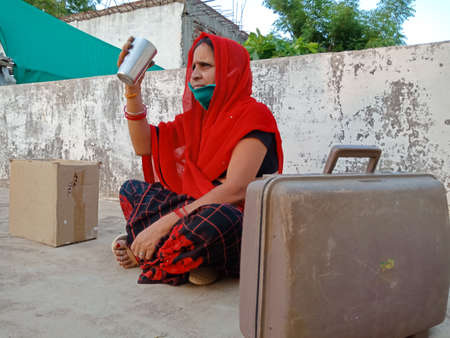DISTRICT KATNI, INDIA - MAY 21, 2020: An indian village lady drinking water sitting with luggage at sky wall background.のeditorial素材