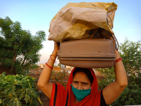 DISTRICT KATNI, INDIA - MAY 21, 2020: An indian village woman wearing face mask for corona virus protection holded lugage on head at sky background.のeditorial素材