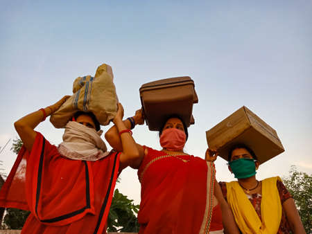 DISTRICT KATNI, INDIA - MAY 21, 2020: group of indian village poor females wearing face mask for corona virus protection holded lugage on head at sky background.のeditorial素材