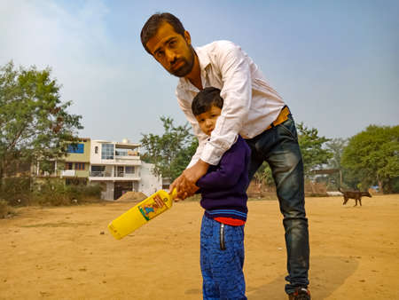 DISTRICT KATNI, INDIA - JANUARY 17, 2020: Indian preschool male teacher teaching to little boy about cricket game together at soil field on sport ground.のeditorial素材