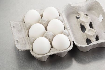 Six white hen eggs in a carton on a scratched metal surface.の写真素材