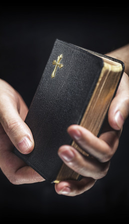 Man holding an old small black bible in his hands. Short depth of field, the sharpness is in the cross.の写真素材