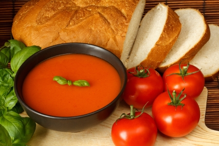 Bowl of tomato soup with basil garnish, in kitchen setting surrounded by ingredientsの写真素材