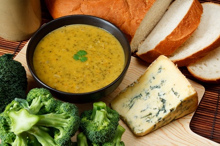 Bowl of broccoli and Stilton soup in kitchen setting surrounded by ingredients, crusty bread and butterの写真素材