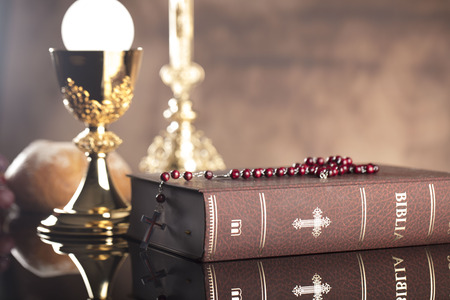 Catholic religion theme. Holy Bible, the cross and gold chalice on wooden table and stone...