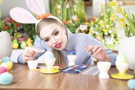 Cute young women with bunny ears painting easter eggs.  Easter background. Bouquets of spring flowers. Easter eggs in baskets.の写真素材