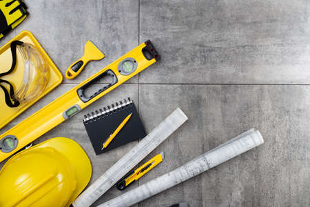 Contractor theme. Tool kit of the contractor: yellow hardhat, libella, hand saw. Plans and notebook on the gray tiles background.の写真素材