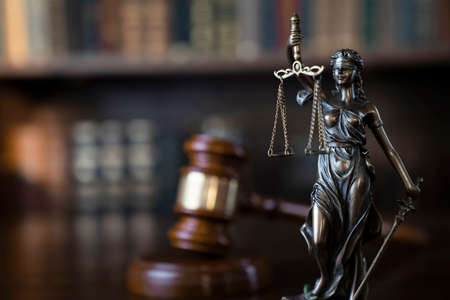 Law theme. Judge chamber. Themis and gavel on brown shining desk. Collection of legal books in the bookshelf.の写真素材