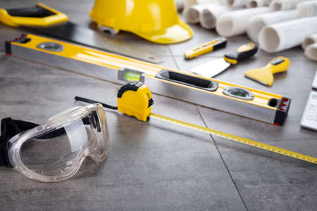 Contractor theme. Tool kit of the contractor: yellow hardhat, libella, hand saw. Plans and notebook on the gray tiles background.の写真素材