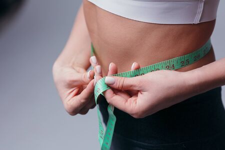 Slender young woman measures her slim waist with a tape measure, closeup in a white tank top and gray leggingsの写真素材