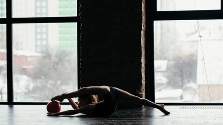 The girl does gymnastics with a red ball near the window. Silhouette on a bright background.の写真素材