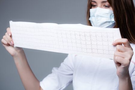 A young doctor girl in medical mask looking cardiogram on white background