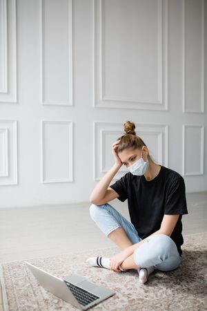 Woman in a t-shirt and medical mask looks puzzled at the laptops screen trying to solve a work problem. Work at homeの写真素材