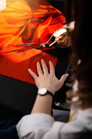 Female tailor's hands cut with scissors a red dense fabric on a cutting table