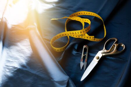 Tailor scissors, tape measure and clipper for thread lies on a dark blue satin fabric shining under the sunの写真素材