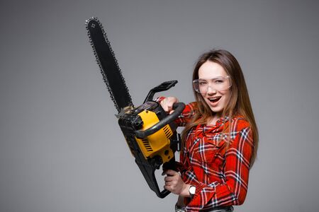 Beautiful sexy girl in red plaid shirt and in transparent glasses holds a chainsaw and crags. Advertising shot for hardware store.の写真素材
