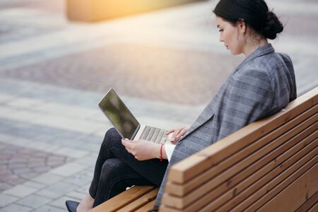 Rear view of a business woman in a jacket sitting on a bench with a laptop on her lap, working outdoors outdoors.の写真素材