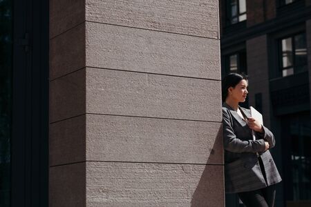 A view from around the corner of a beautiful girl in a jacket looking at people and holding a laptopの写真素材