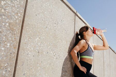 Outdoor portrait of a beautiful girl in a sports top drinking water from a sports bottle after...