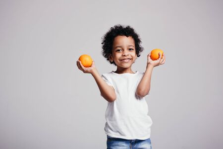 A little Maracanian boy holds two oranges in two hands and smiles happily while in the studio on a light backgroundの写真素材