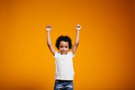 A curly-skinned African-American boy in a white T-shirt and jeans joyfully extended his arms up against a orange backgroundの写真素材