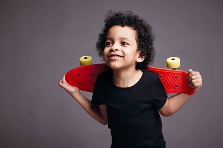 Little black boy in a black T-shirt holds a red longboard behind his back and smiles in the studioの写真素材